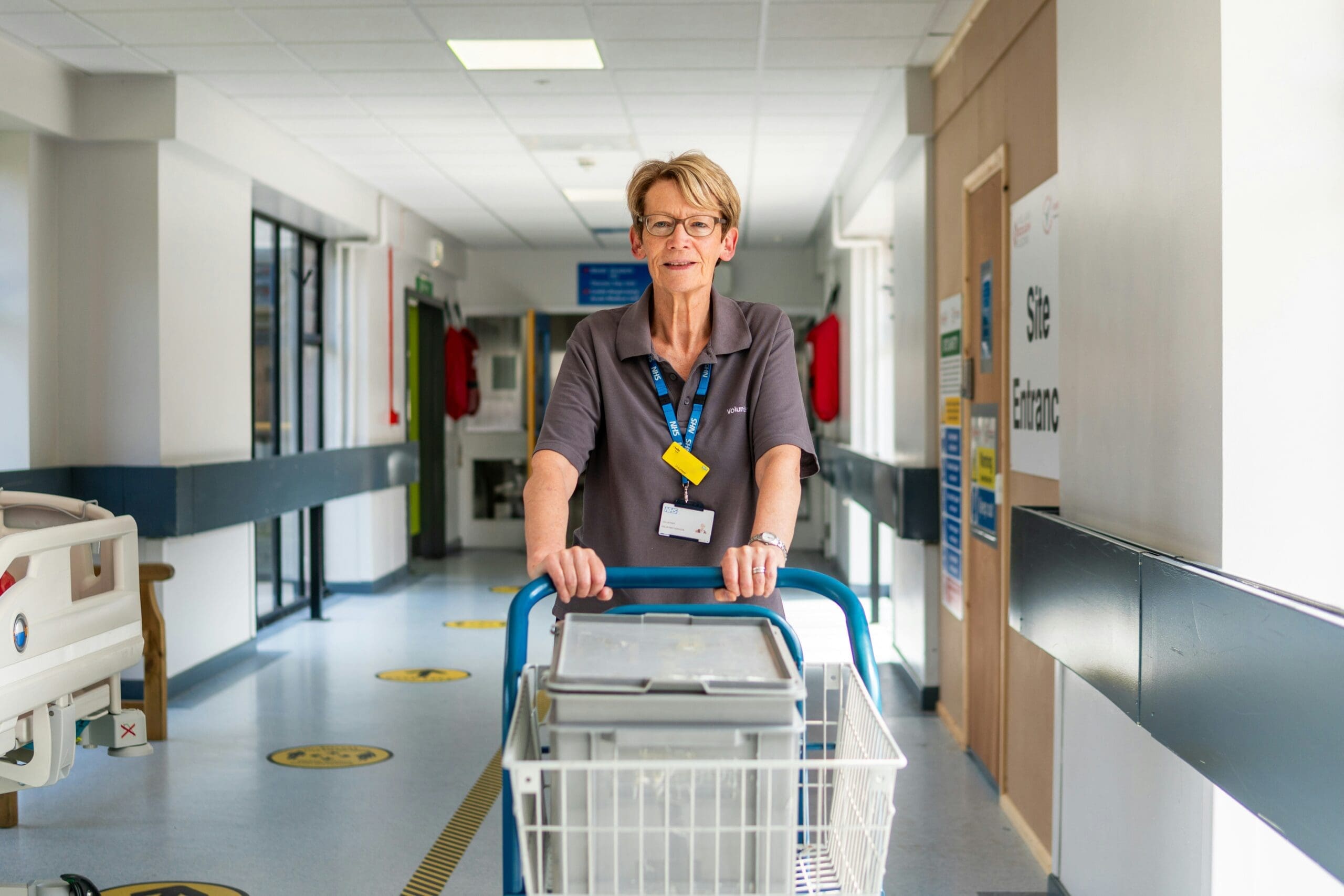 An NHS hospital volunteer wheeling a trolley along a busy hospital corridor, with patients and staff visible in the background