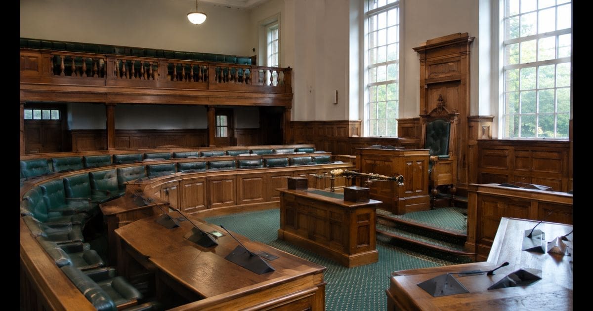 Empty UK council chamber interior with horseshoe wooden bench seating, oak panelling and soft daylight from tall windows, no people